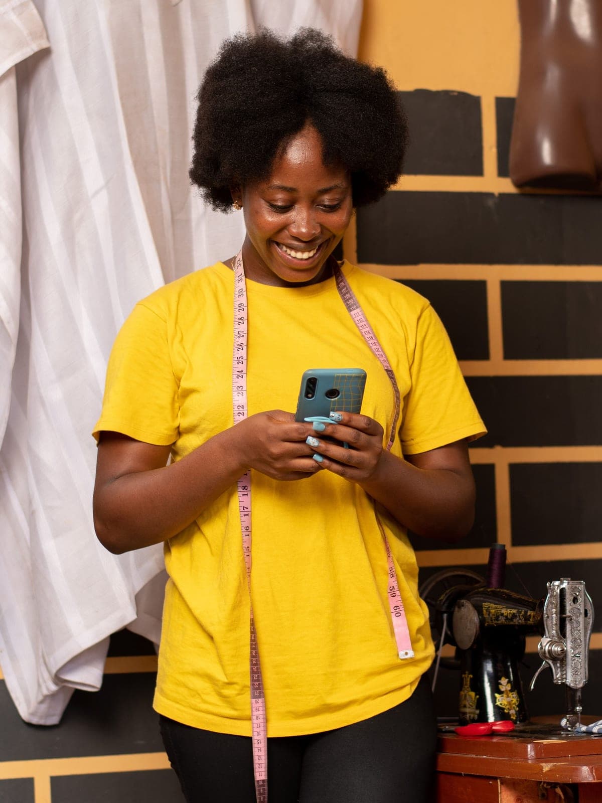 A tailor smiling at a phone in a workshop, tape measure around the neck and a sewing machine alongside